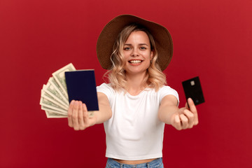 Happy girl with curly blond hair in a white t-shirt and a sundown hat on a red background. Model holds pasport with cash and a bank card.
