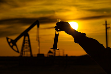 A man holding a flask of oil. Oil rigs in the background. Oil production in Russia. Sunset.