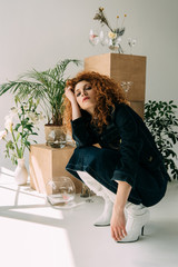 trendy redhead girl sitting and posing near wooden boxes, glasses and plants on grey