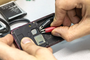 Elderly and young men repair a mobile phone together. They work in a repair shop. They use different tools. They are repair specialists