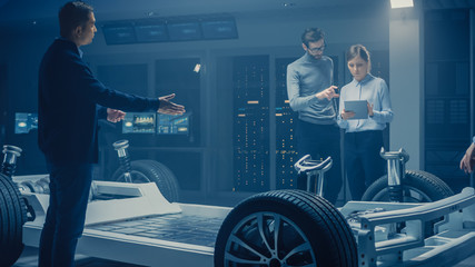 Automobile Engineers Discussing and Designing Electric Car Chassis Platform, Using Tablet Computers with 3D CAD Software. In Automotive Innovation Facility Vehicle Frame with Wheels Engine and Battery