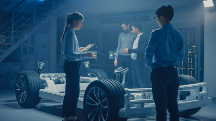 Automobile Engineers Discussing and Designing Electric Car Chassis Platform, Using Tablet Computers with 3D CAD Software. In Automotive Innovation Facility Vehicle Frame with Wheels Engine and Battery