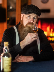 Bearded Scottish man enjoys drinking old whiskey in living room with fireplace