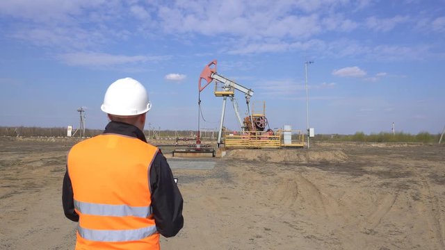 A male inspector-operator in a signal vest and a white helmet records the readings about the pumping unit oil, gas and combustible fuel, industry, copy space, pumpjack