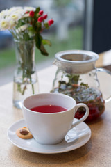 Cup of red herbal tea, cookie, teapot, vase with flowers on the table. Close-up, copy space, vertical format