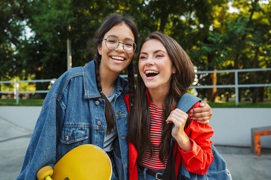 Image of two caucasian girls laughing and hugging together while holding skateboard in skate park