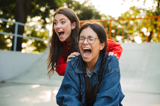 Image of two delighted girls screaming and riding skateboard together in skate park