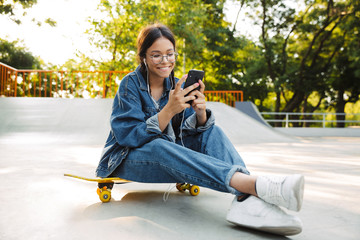 Image of beautiful girl using smartphone and earphones while sitting on skateboard in skate park