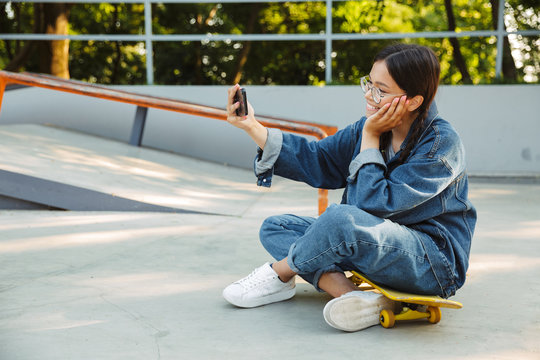 Image of pretty girl taking selfie photo on smartphone while sitting on skateboard in skate park