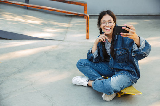 Image of cute girl taking selfie photo on smartphone while sitting on skateboard in skate park