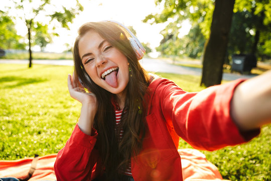 Image of funny girl with tongue sticking out using headphones and taking selfie photo while sitting in green park