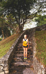 Blonde in denim shorts standing in the park on the stone steps. Wearing white jersey, orange backpack. Hands with tattoos. Asia travel