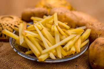 Raw cut french fries in a transparent glass plate along with raw potato with it on jute bag's surface.