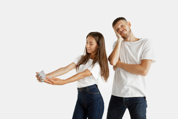 Beautiful young couple's portrait isolated on white studio background. Facial expression, human emotions, advertising concept. Woman making selfie or video for vlog, man is bored, doesn't want to do.