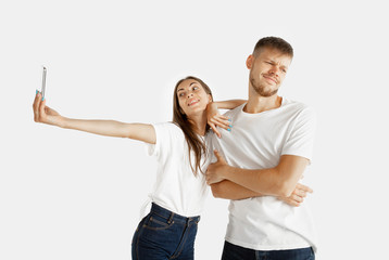 Beautiful young couple's portrait isolated on white studio background. Facial expression, human emotions, advertising concept. Woman making selfie or video for vlog, man is bored, doesn't want to do.