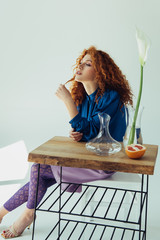 stylish redhead girl posing at table with calla flower and vases on grey