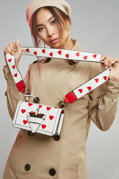 Three quarter portrait of Asian lady with dark hair, wearing trench coat and beige beret. The woman is tilting her head while stretching the bag's strap. The clatch is adorned with heart shape print.