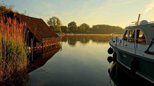 ruhige Morgenlandschaft in der Mecklenburger Seenplatte mit Hausboot und reetgedecktem Bootshaus