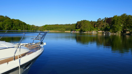 Naklejka na ściany i meble weißes Boot fährt auf stillem See umrahmt von grünem Wald in der Mecklenburger Seenplatte