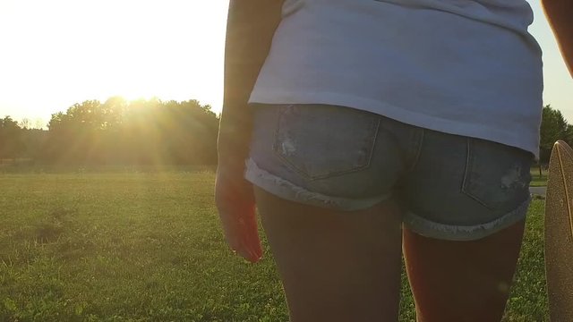 SLOW MOTION Attractive sexy young woman legs wearing jeans shorts holding a skateboard, close-up shot from behind with camera circling subject, lens flare at sunset