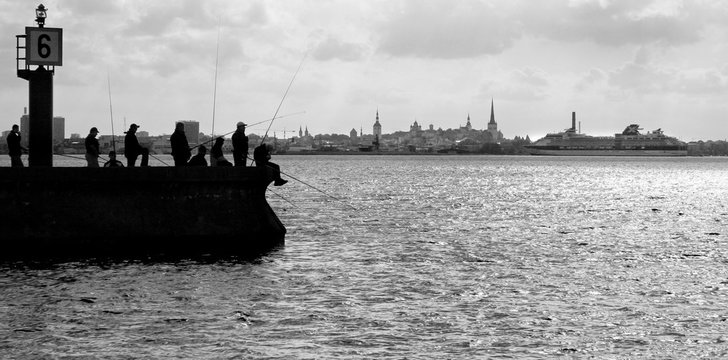Silhouette photo fishermen in backlight fishing on the mole on the background of Tallinn. A big ferry approaches the city. A lot of fishing rods. Black and white genre photo. On the pier lighthouse