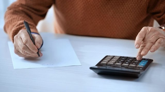 cropped view of senior man writing on paper and counting while using calculator 