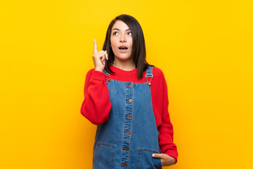 Young Mexican woman with overalls over yellow wall thinking an idea pointing the finger up