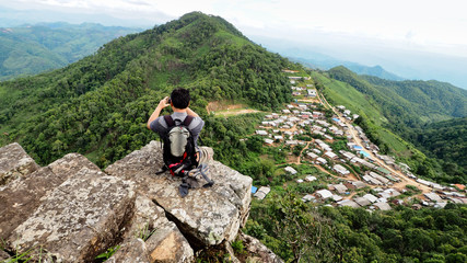 Landscape Photography of A Man Sitting on A Cliff and Taking Photos of The Rural Village with Mobile or Smart Phone with Hills / Mountains, Blue Sky and White Clouds in The Background.