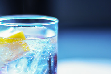 A piece of glass with a cold refreshing carbonated drink with ice and lemon close-up on a gray-blue background. Gas bubbles in cold water through the glass of a glass.Copy Space