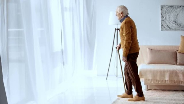 senior man with grey hair holding walking stick while walking in living room 