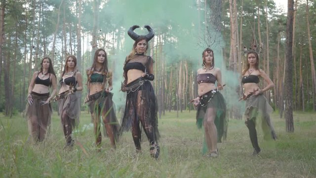 Group of women dancers with make-up and in mystical fabulous costumes dancing in color smoke. Forest fairies, dryads have fun among the trees. Performance of dancers outdoors.