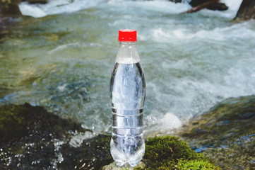 A plastic bottle with a red cap with fresh drinking water against a background of green forest and a mountain river stands on a stone with moss. The concept of pure natural mountain water