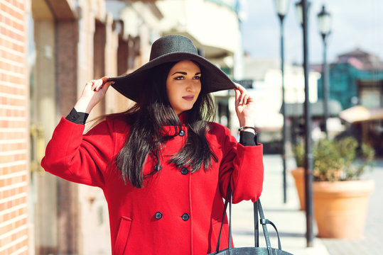 Young pretty trendy girl posing at the city in Europe. Fashion woman portrait outdoors. Autumn women fashion. Girl holding retro black hat. Woman wearing red trendy coat