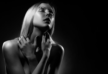 Beautiful blonde is crying, covering her topless big breasts with her hands. Tears on her face. Avant-garde makeup, eyebrows made of foil. Dark background. Conceptual artistic black and white photo