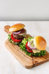 Two homemade fast food burgers classic hamburger or cheeseburger with beef, salad, cheese and tomato served on wooden cutting board on white marble kitchen table. Copy space.