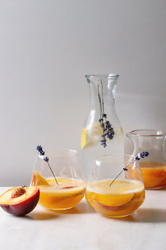 Peach cold lemonade or Bellini cocktail with lavender flowers served in two glasses with pitcher of pesh juice and lemon soda water on white marble table. Homemade summer drink. Copy space