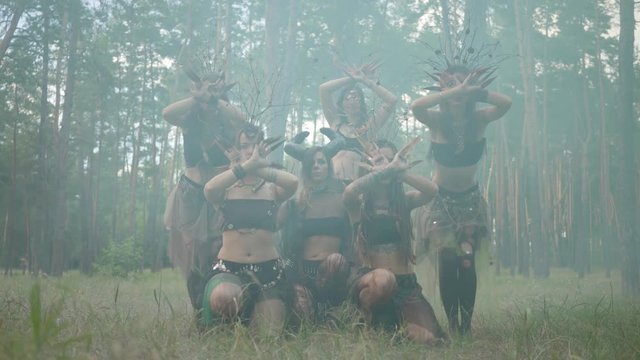 Group of women dancers with make-up and in mystical fabulous costumes dancing groovy dance in color smoke. Forest fairies, dryads have fun among the trees. Performance of dancers outdoors.