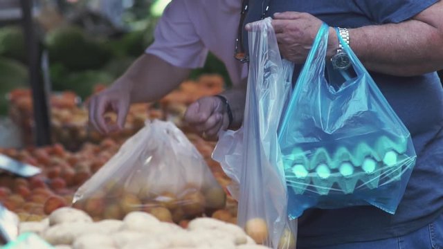Male customer choosing peaches filling a plastic bag while carrying a blue plastic shopping bag filled with eggs at a farmers street market.