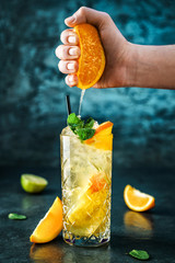 Fresh lemonade with mint, ginger, orange and ice in glass jar on the dark blue background. Man hand squeezes orange juice from orange in glass with splash and drops. Summer cold drink and cocktail