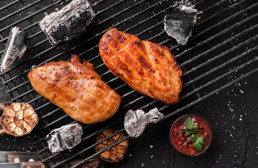 Grilled chicken breast meat with garlic over the coals on a barbecue, dark background with light of fire. Top view