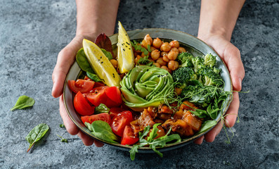 Hands holding healthy superbowl or Buddha bowl with salad, baked sweet potatoes, chickpeas, broccoli, hummus, avocado, sprouts on light blue background. Healthy vegan food, clean eating, dieting