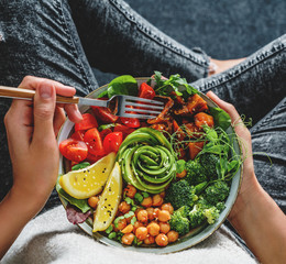 Woman in jeans holding Buddha bowl with salad, baked sweet potatoes, chickpeas, broccoli, greens,...