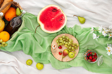 Summer picnic in the meadow on the green grass. Fruit basket, juice and bottled wine, watermelon, strawberries in a glass jar and baguettes of bread. White tablecloth and a bouquet of field daisies.