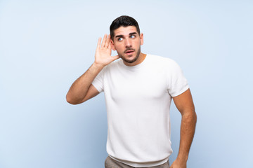 Caucasian handsome man over isolated blue background listening to something by putting hand on the...