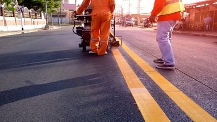 Selective focus of road workers with thermoplastic spray road marking machine working to paint yellow line on asphalt road surface with flare light in evening time, construction concept - obrazy, fototapety, plakaty