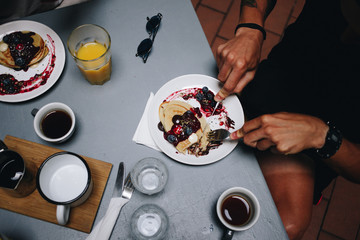 Man with arm tattoos enjoys fancy breakfast at cute popular cafe downtown, cuts organic whole wheat pancakes with berries at table served with orange juice and filter coffee. Food blogger style