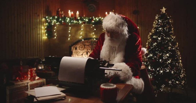 Senior bearded man in santa claus clothing is working on letter at his typewriter in christmas decorated room - christmas spirit, holidays and celebrations concept 4k footage