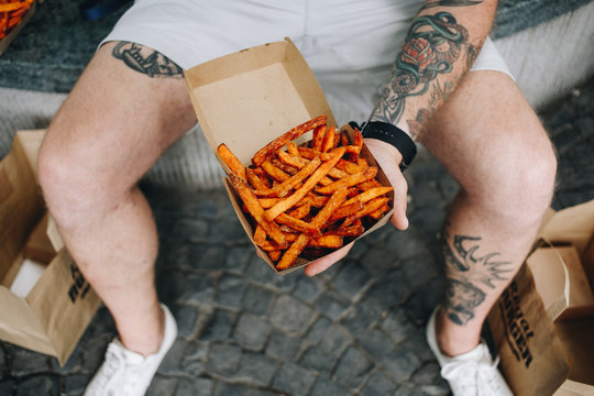 Cinematic photo of hipster millennial man with body tattoos hold open take away box of delicious crispy and organic sweet potato fries. Street food lovers and blogger lifestyle, summer snacks
