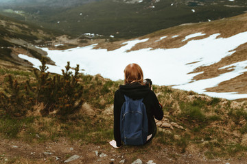 A young blonde travels with a blue backpack, sits on top of a mountain and enjoys green mountain scenery. Back view