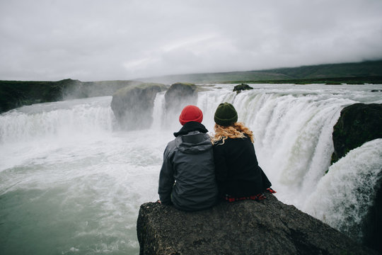 Couple of travellers in winter warm clothing sit on steep dangerous cliff next to waterfall, watch falling water, calm and astonished by power of nature. Relationship goals, explore more